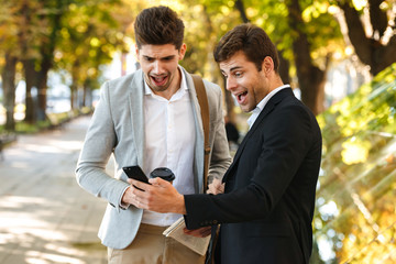 Photo of joyous businessmen in suits using smartphone while walking outdoor through green park with takeaway coffee, during sunny day