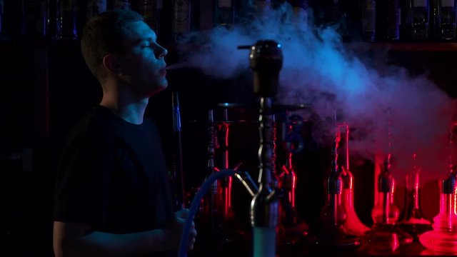 Confident man resting in lounge restaurant on against black background, smoking hookah. A young man smokes a hookah in a hookah bar on the background of a bar with hookahs in the dark, slow motion.