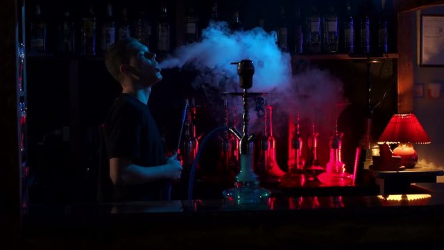 Silhouette of a young man in a hookah bar against the background of a bar with various colored hookahs, he smokes fruit tobacco and blows a lot of smoke. Slow motion. A man smokes a hookah.