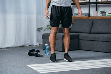 cropped view of man with tattoo on hand standing near fitness mat in living room