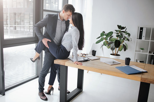 Sexual and intimate picture of couple at work. She sit on table. He hold her leg in sexual pose. They keep faces close to each other.