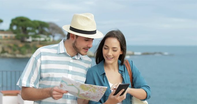Happy couple of tourists walking comparing smart phone content and paper map on the beach