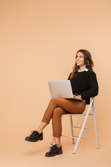 Beautiful amazing young woman posing isolated sitting on stool using laptop computer.