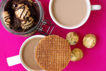 Two cups with coffee, caramel waffles, jar with cookies and sweets wrapped in golden foil, top view, pink background