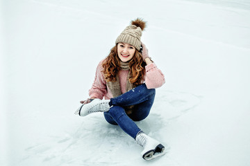 Beautiful girl in warm clothes sitting on ice rink after falling and laughing