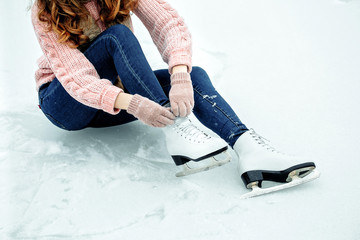 Girl tying shoelaces on ice skates before skating on the ice rink, hands in red knitted gloves. View from top.