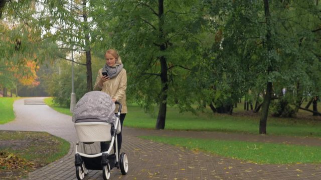 Mother having an walk with baby in the park on autumn day. She pushing pram and using smart phone