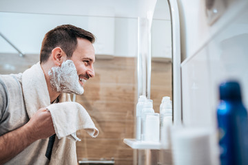 Portrait of a handsome man with shaving cream on his face and towel while standing in the bathroom, looking at mirror. - Powered by Adobe