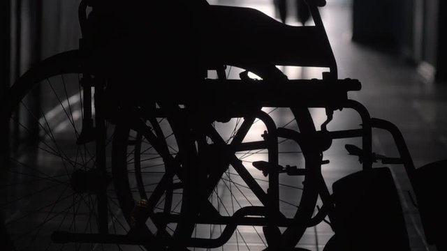Tilt up shot of silhouette of woman with stick limping to wheelchair along hallway of hospital