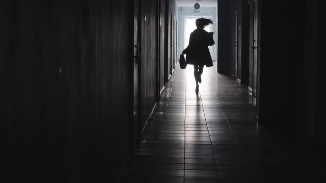 Tilt up shot of silhouette of businesswoman with bag and documents running towards the camera along hallway in office center