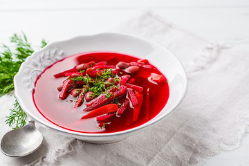 Vegetarian beet soup with beans and vegetables in white plate on white wooden background.