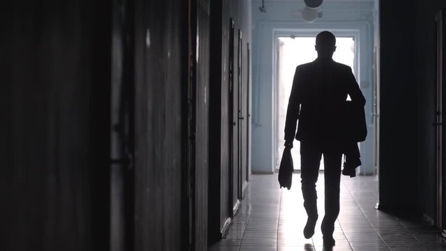 Silhouette of businessman wearing formal suit and carrying coat and briefcase walking towards the camera along hallway in office center