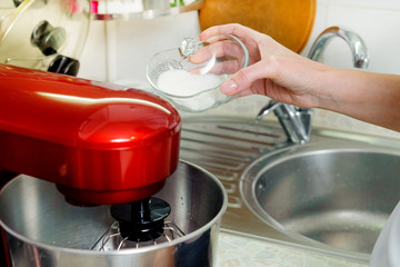 A woman is cooking in her kitchen, about to bake a cake. Woman kneads dough in red food processor, adds sugar to dough