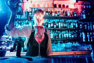 Glass of fiery cocktail on the bar counter against the background of bartenders hands with fire. Barman day concept