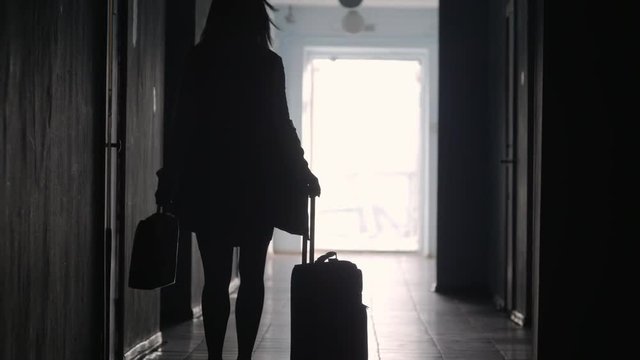 Rear view of full length silhouette of businesswoman carrying briefcase and pulling luggage while walking along hallway in hotel