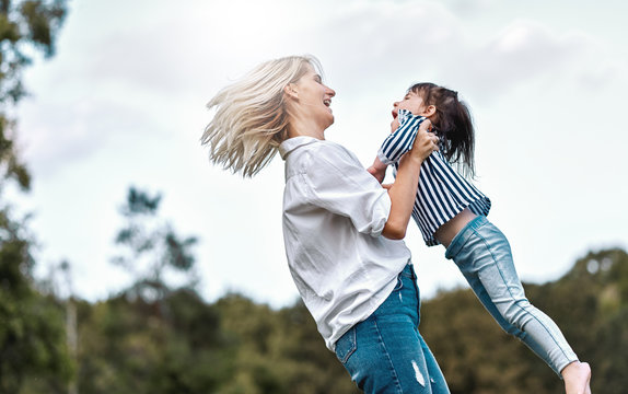 Side view image of happy little daughter playing with her smiling mother in the park. Loving woman and her little kid girl spinning together. Mom and child has fun outdoors. Mother's day