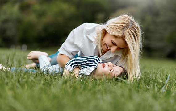 Happy little daughter lying on the green grass and playing with her smiling mother in the park. Loving woman and her little kid girl enjoying the holiday. Mom and child has fun outdoors. Mother's day