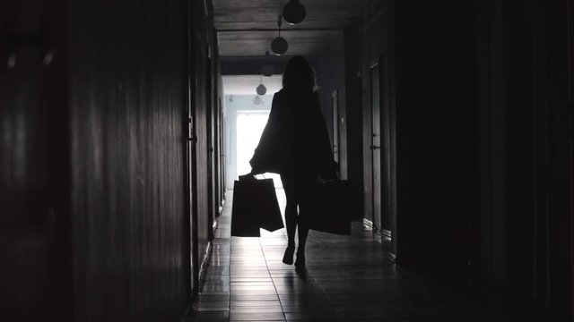 Rear view full length silhouette of unrecognizable woman wearing high heels and walking with shopping bags along hallway in hotel 