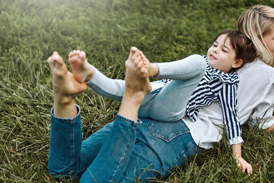Horizontal image of beautiful woman lying and playing with her cute little girl on the green grass outdoors. Happy child and mother spend time together in the park. Mother's day concept