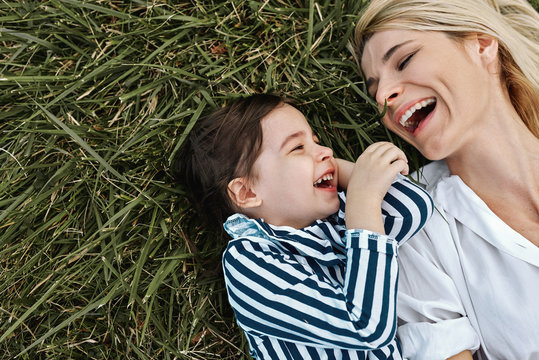 Closeup overhead image of happy beautiful woman laughing and playing with her cute little girl lying on green grass in the park. Mother and daughter has fun together. Mother's day