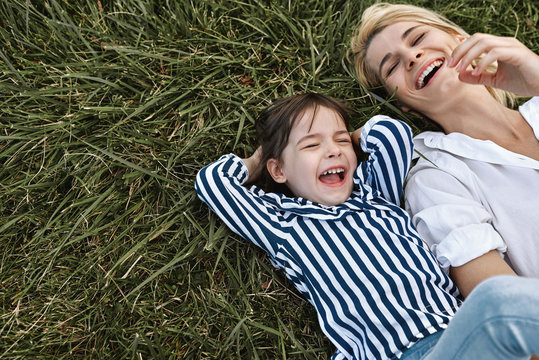 Horizontal overhead image of happy beautiful woman laughing and playing with her cute little girl lying on green grass in the park. Mother and daughter has fun together. Mother's day