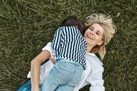 Top view image of happy beautiful woman smiling and playing with her cute little girl lying on green grass in the park. Mother and daughter has fun together. Mother's day