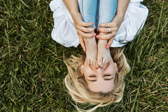 Overhead image of cheerful blonde woman playing with her cute little child, lying on green grass outdoor. Happy mother and daughter spend time together in the park. Mom and kid has fun. Mother's day