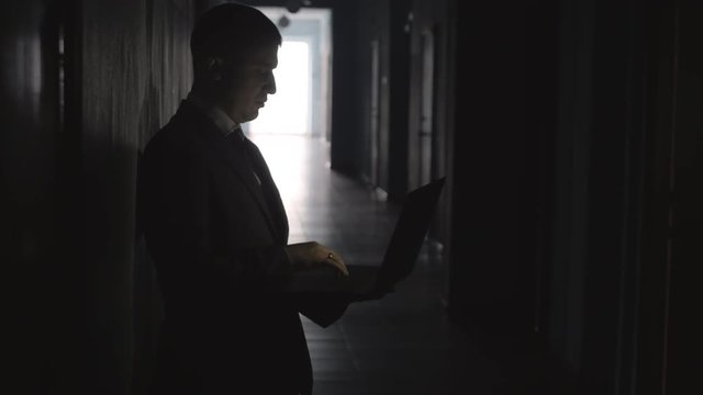 Tilt up of silhouette of businessman in formal suit standing in dark hallway and typing on laptop
