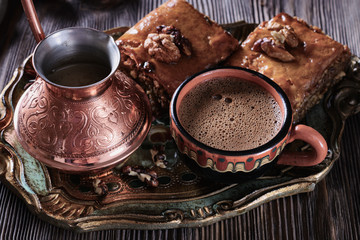 A cup of freshly brewed coffee next to a cezve and baklava on a wooden vintage tray and on a wooden background, closeup
