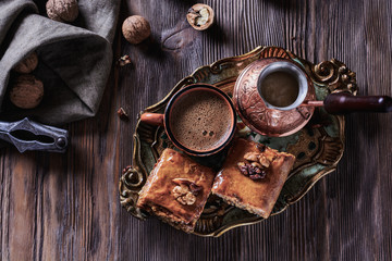 A cup of freshly brewed coffee next to a cezve and baklava on a wooden vintage tray and on a wooden background next to nuts, top view, closeup