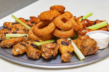 Bar Food:  Chicken wing platter with onion rings and celery.  Container of white dipping sauce on white board.  Side view close up.
