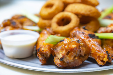 Bar Food:  Chicken wing platter up extreme close.  White dipping sauce on the side.  Onion rings blurred in the background.