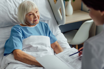 doctor holding diagnosis and consulting senior woman with grey hair lying in hospital bed