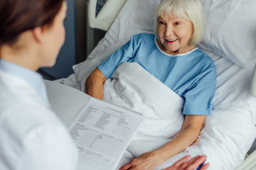 doctor holding diagnosis and consulting smiling senior woman lying in hospital bed