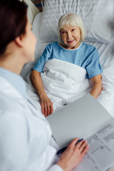 female doctor sitting on bed and holding diagnosis while senior woman lying in bed in hospital