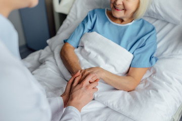 cropped view of female doctor holding hands with senior woman lying in bed in hospital
