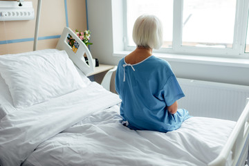 Fototapeta premium back view of senior woman sitting on bed in hospital ward