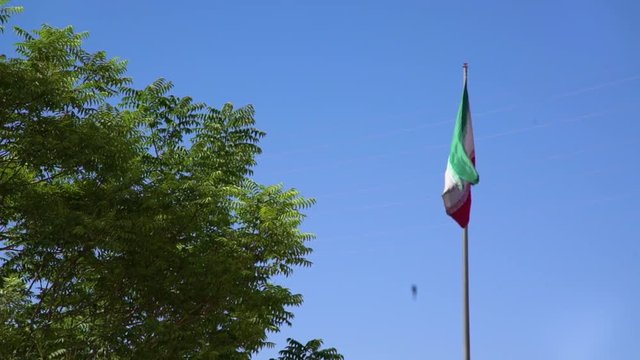 Mid shot of the flag of Iran flapping in the wind on a tall pole next to a tree with blue sky