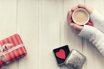 Love concept. Female hands holding a cup of coffee, red heart in a box on a wooden table. Top view.