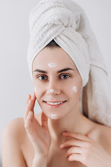 Young happy woman wrapped with bath towel applying  cream to her face for skin care. Skin care...