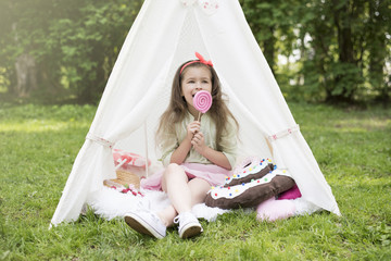 Little girl playing in a tent, children's house wigwam in park. Happy mothers day. Child with big donuts, lollipop and straw hat. Summer, happy childhood concept. Cute emotions girl. © MartaKlos