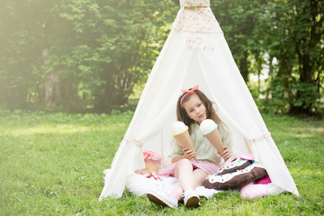 Little girl playing in a tent, children's house wigwam in park. Happy mothers day. Child with big donuts and ice cream and straw hat. Summer, happy childhood concept. Cute emotions girl. © MartaKlos