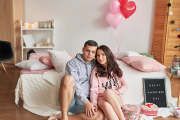 Young couple in love at home on bed celebrates Valentine's Day on February 14, bouquet of red roses and heart-shaped balloons decorate  room. Saint Valentine Postcard template. Love story. 