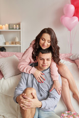Young couple in love at home on bed celebrates Valentine's Day on February 14, bouquet of red roses and heart-shaped balloons decorate  room. Saint Valentine Postcard template. Love story. 