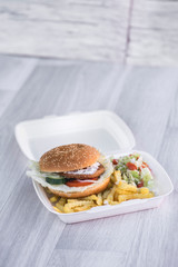 tasty delicious hamburger in box with French fries and vegetable, grey background, wood pattern, light picture, close up of meal, unhealthy fast food option for eating