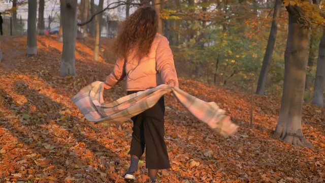 Portrait of young caucasian curly-haired woman walking and spinning around happily in autumnal park.