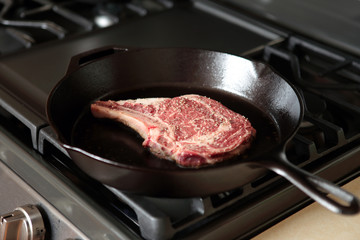 Rib eye steak cooking in a cast iron pan on a natural gas stove top.
