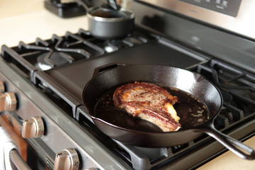 Rib eye steak frying in a cast iron pan on a natural gas stove top.