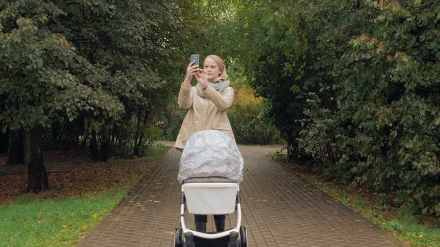 A woman with a baby carriage is walking along a paved walkway in an autumn park. She stops to take a picture of surroundings on her smartphone and then continues her way