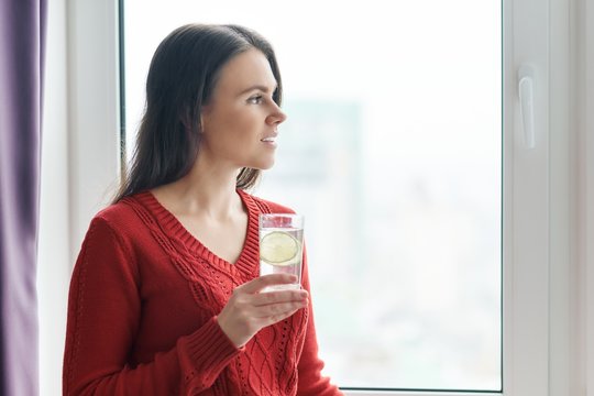 Young Beautiful Woman In Red Sweater With Glass Of Water With Lime, Woman Stands Near Window In Skyscraper On Cloudy Day. Healthy Drink, Natural Antioxidant, Vitamins In Urban Life